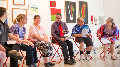 Artists sitting on red fold out chair in a gallery delivering artist talks with their artworks hagning on the walls in the background.