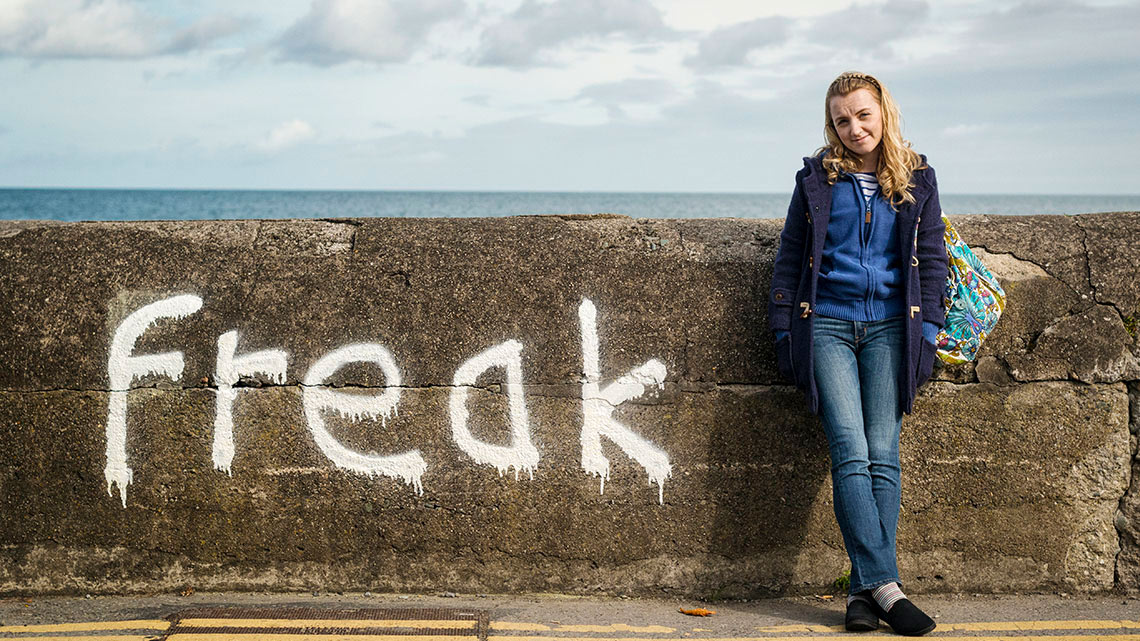 Girl standing in front of a concrete wall with 'freak' spray painted on it