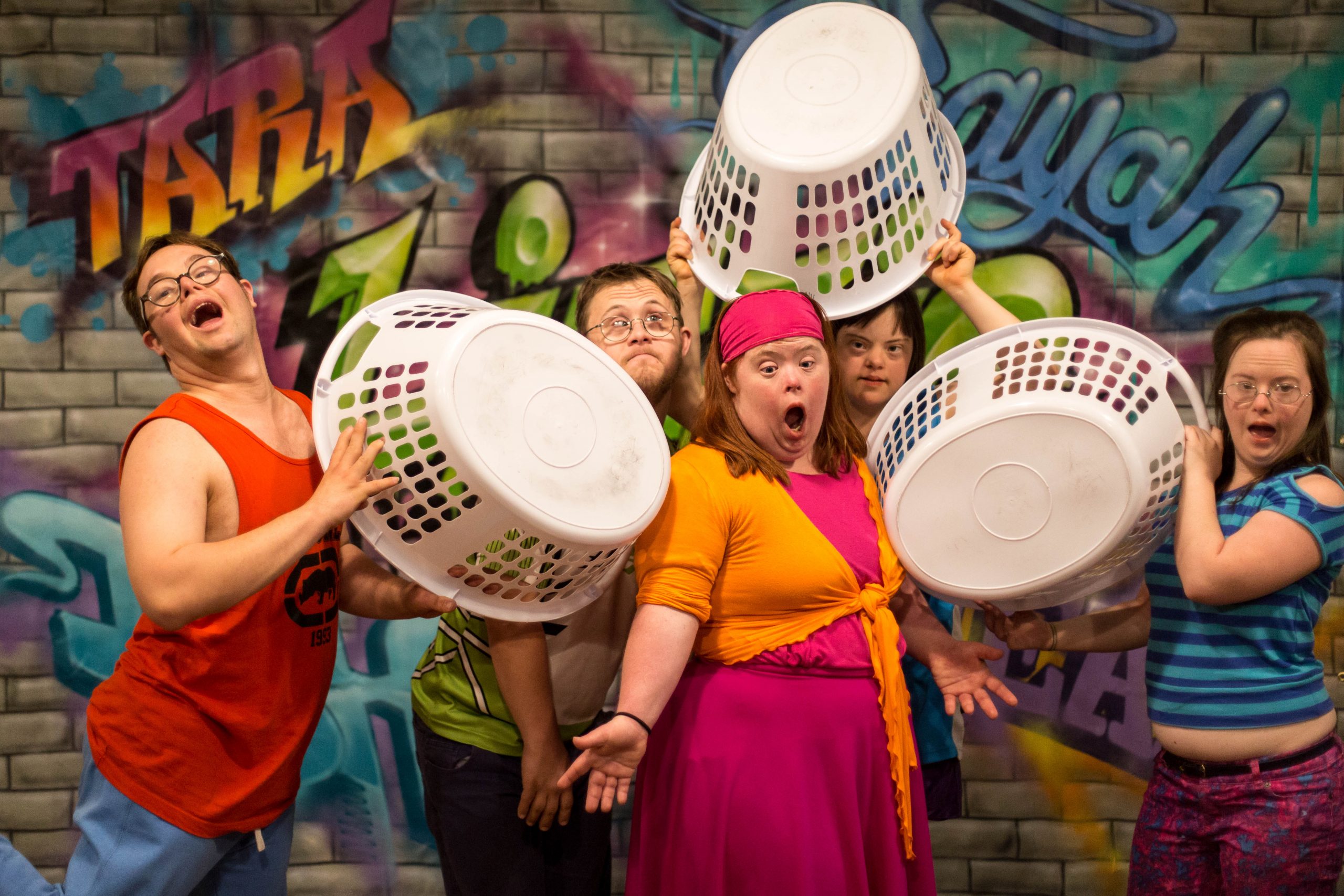 Five actors are on stage, wearing brightly coloured clothes and against a backdrop of a wall covered in graffiti.Three hold white laundry baskets and three appear to be singing.