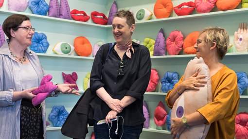 Three women stand in the Bella Room, which is covered in light green shelves filled with colourful cushioned artworks in the colourful shapes of mouths, eyes, noses and other objects.