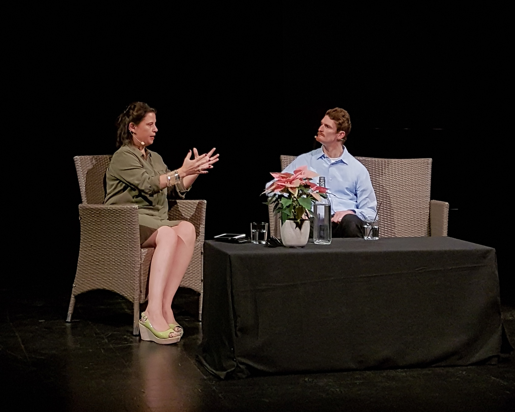 Michelle Silby sits on a chair. She wears a green dress and is gesturing with her hands while facing Dan Daw. Dan sits in a chair facing Michelle and is looking at her. The background is dark