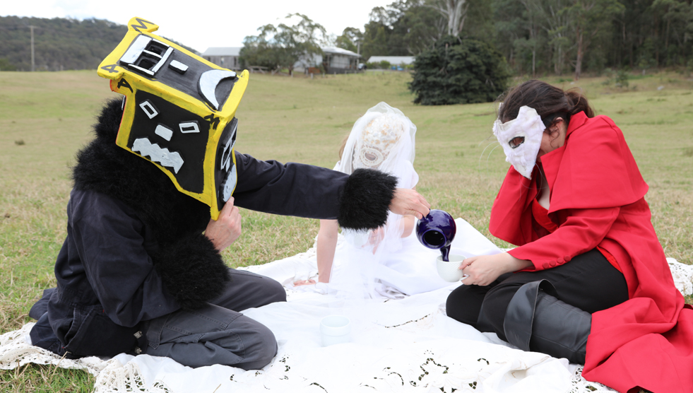 Bundanon Trust Residency: Three people wearing masks are sitting in fancy dress on a picnic mat. One is pouring tea for another