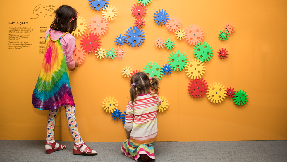 Survey: Two young girls are facing a yellow wall hung with colourful gears. Photo by Marinco Kojdanovski