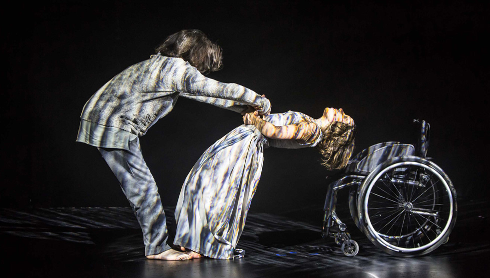 Days: A male and female performer are on stage. She is leaning backwards. He is holding onto her. There is a wheelchair behind her.