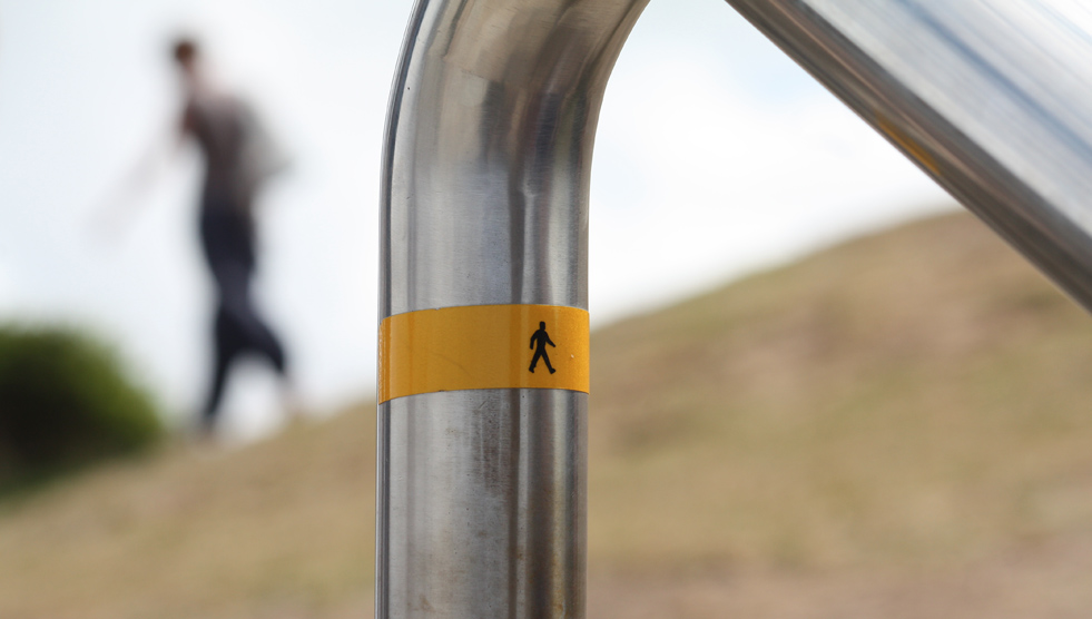 A steel railing is in the foreground with a hiking symbol. A walker is in the background