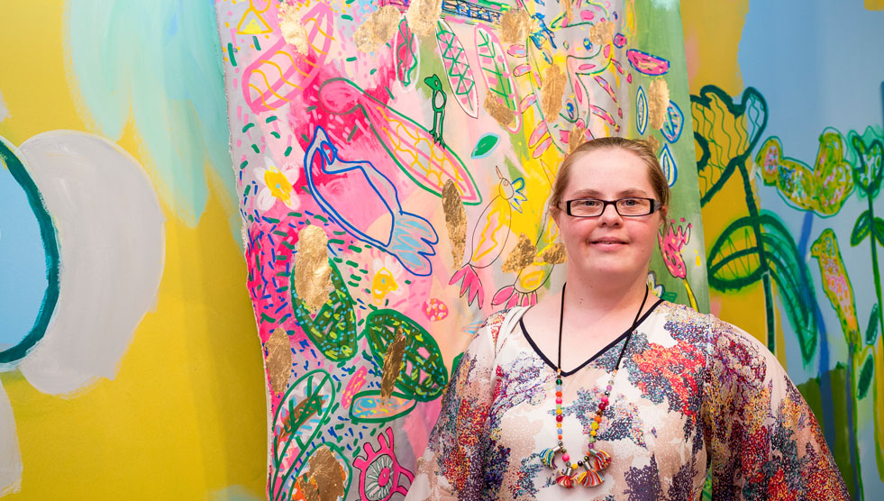National Arts and Disability Strategy: A woman Emily Crockford is standing in front of a large and colourful artwork
