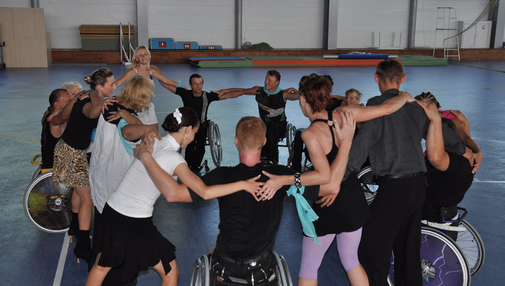 A group of para (wheelchair) dance sport participants are in a circle and are connected with their arms