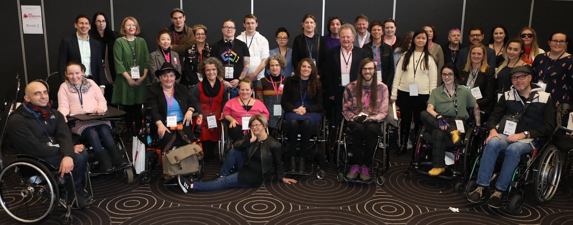 Image is a large group of people sitting on carpet and standing against a wall. Some are in wheelchair, some are in chair, some are standing.