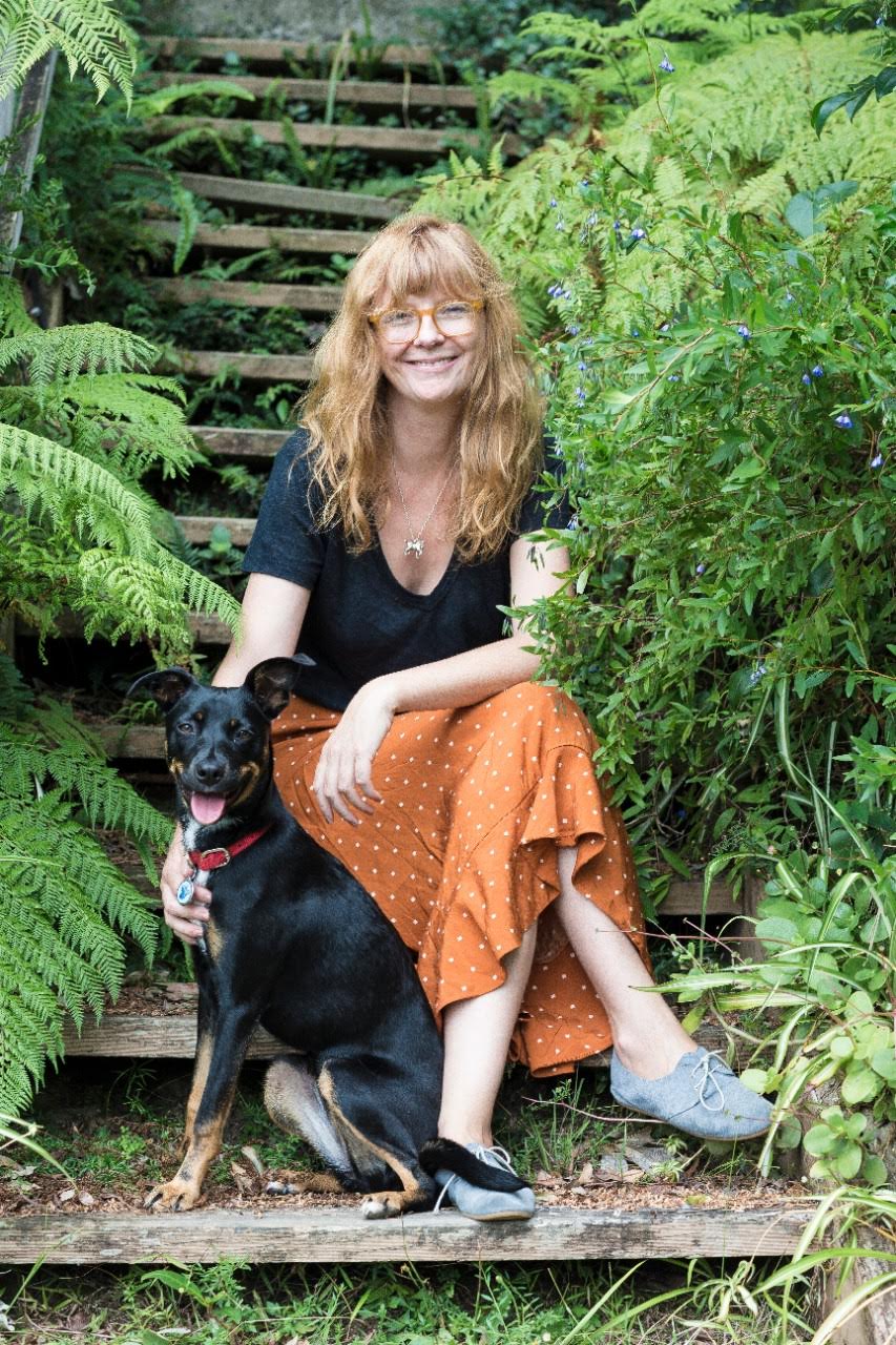 A woman is sitting on stairs in a garden. She's wearing a black shirt and orange skirt. A black dog is sitting next to her 