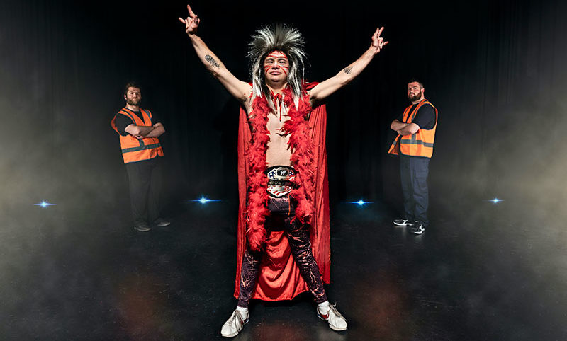 A young man is standing on stage with his arms raised. He is wearing a red cloak and a wig. There are two men on either side of him wearing fluro work vests.