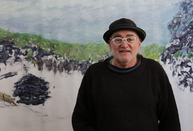 A man is standing in front of a painting. He is wearing a black hat, black jumper and glasses