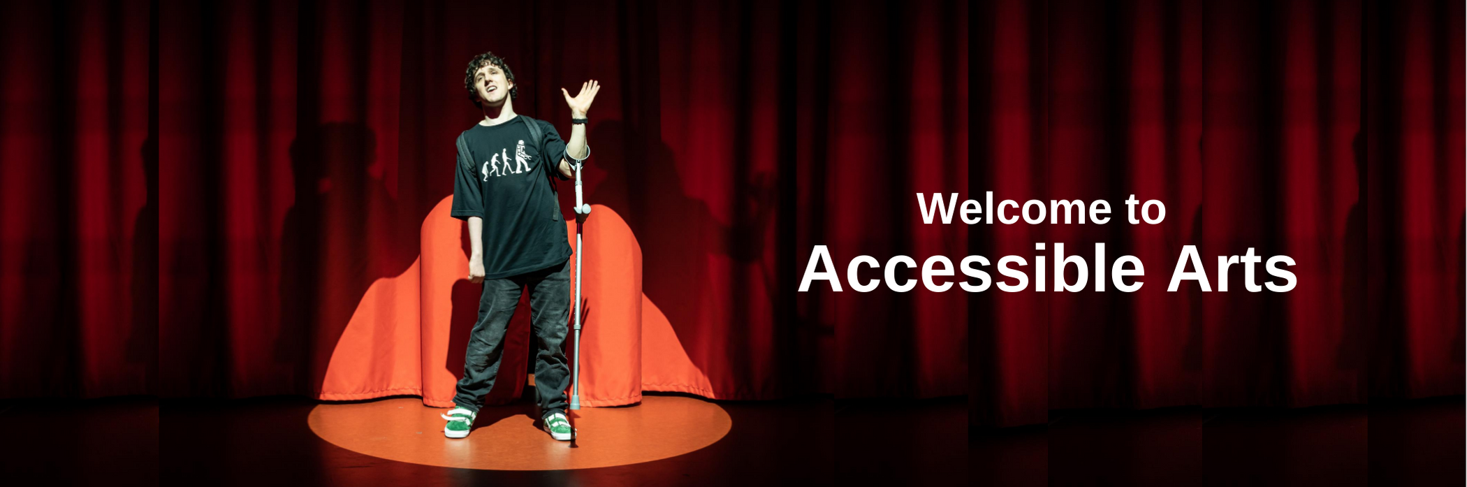 A young man is standing on a stage in front of a red curtain. He is illuminated by a spotlight. His right arm is raised and a walking brace is hanging from his arm. On his right are the words WELCOME TO ACCESSIBLE ARTS.