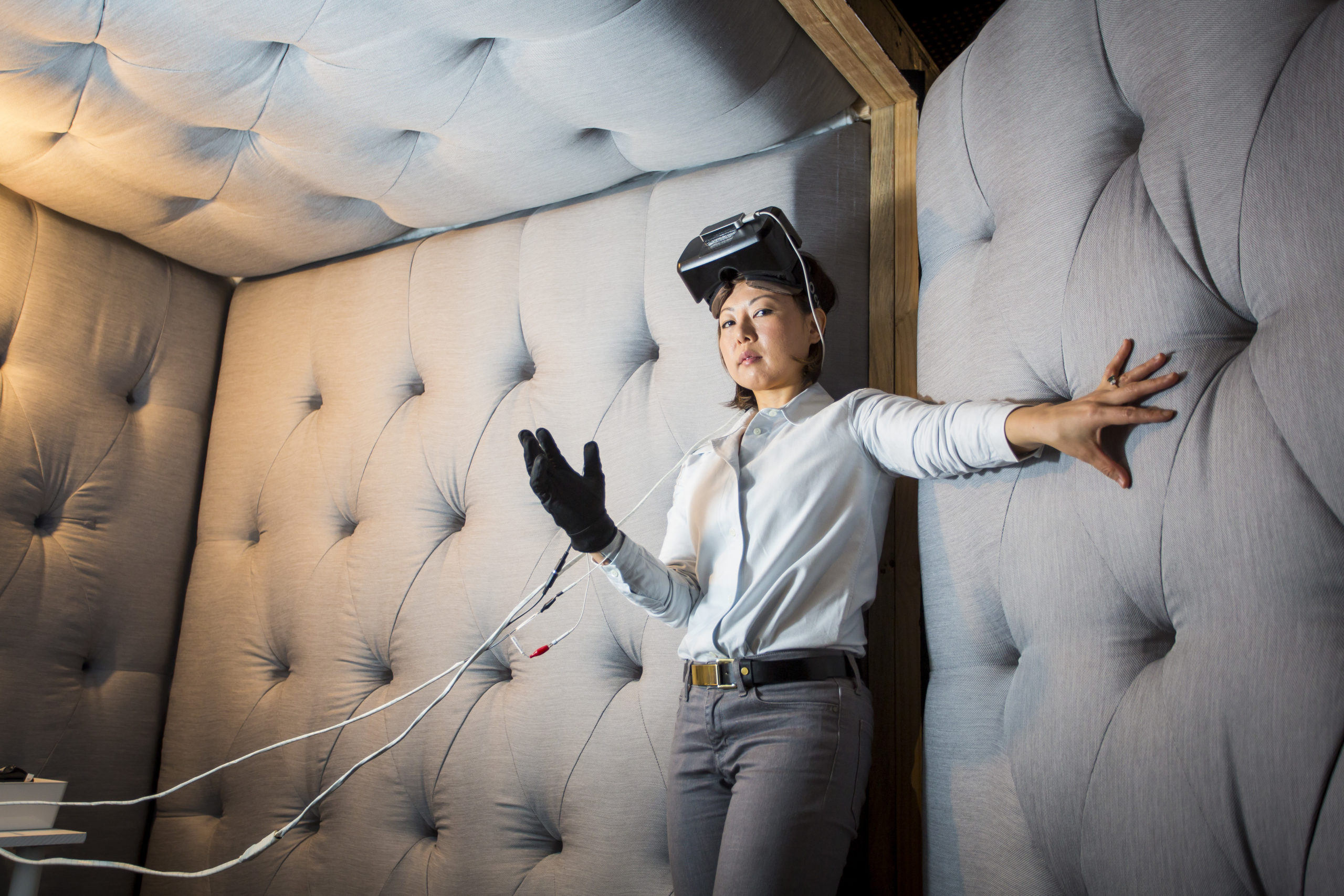 A woman in a grey padded room. her right arm is stretched out and her left hand is in a black glove connected to wires. she is wearing a VR headset on her head