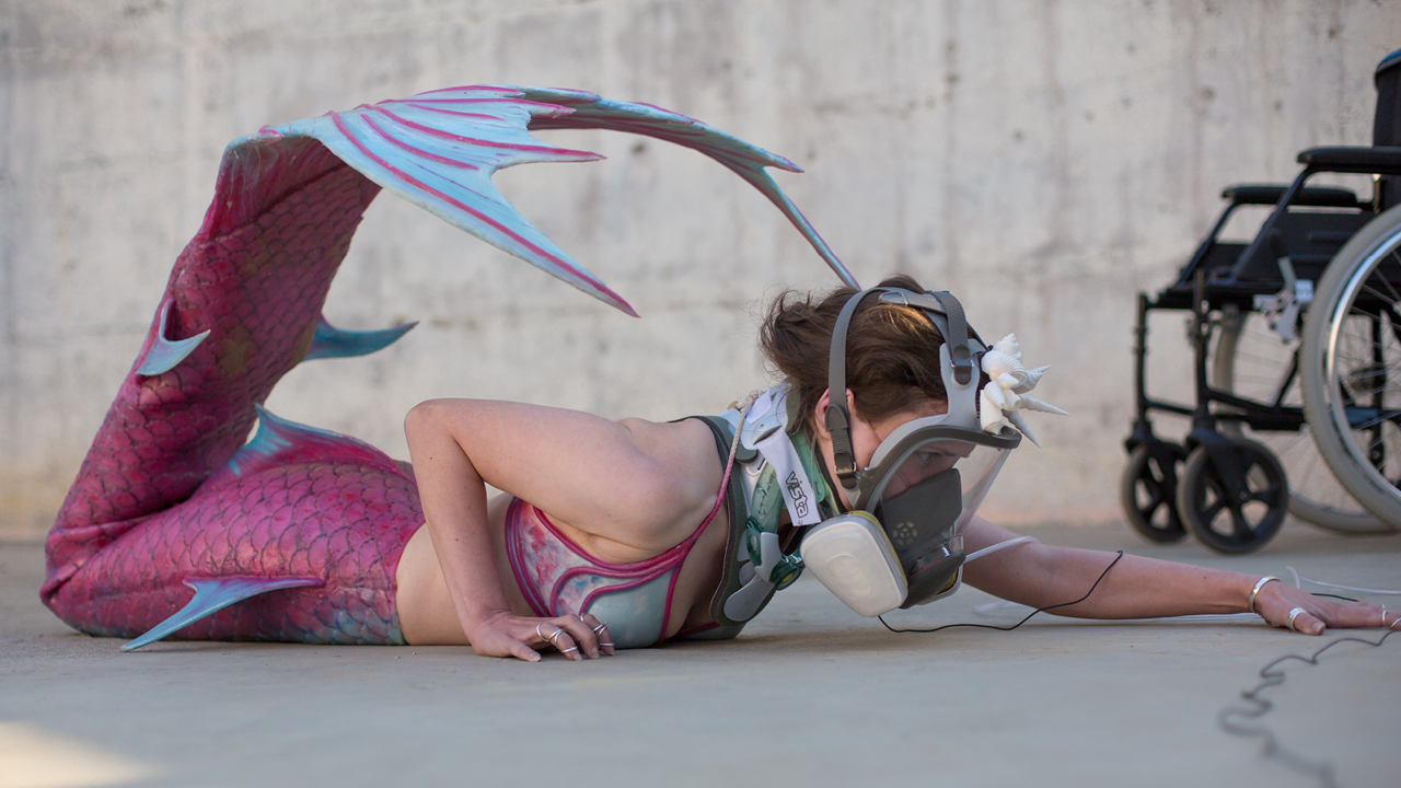 A woman wearing a mermaid costume and gas mask lying on the ground with a wheelchair in the background