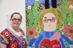 A woman wearing a colourful top and glasses is stadding next to a colourful painting featuirng a woman in blue dress standing in a garden.