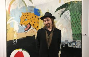 A man is standing in front of a painting. He is wearing a dark suit and hat and has long hair and a beard.