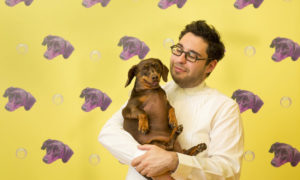 A man is holding a dog. the Man is wearing a white shirt. He has short dark hair and is wearing dark rimmed glasses. They are in front of a yellow wall with repeating pictures of purple dogs.