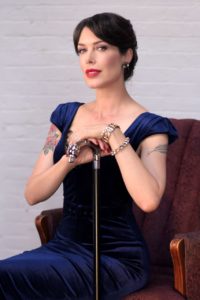 A woman with dark pulled back hair is wearing a dark blue dress and sitting in a red chair. Her hands are crossed on top of a walking stick which stands in front of her.