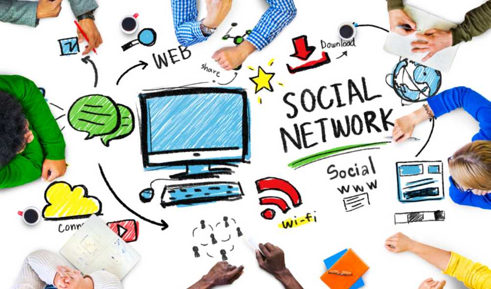 Several people are sitting around table but you can only see their arms. The table is covered in various colourful illustrations of components of digital communications such as computers, mobile phones, and icons.